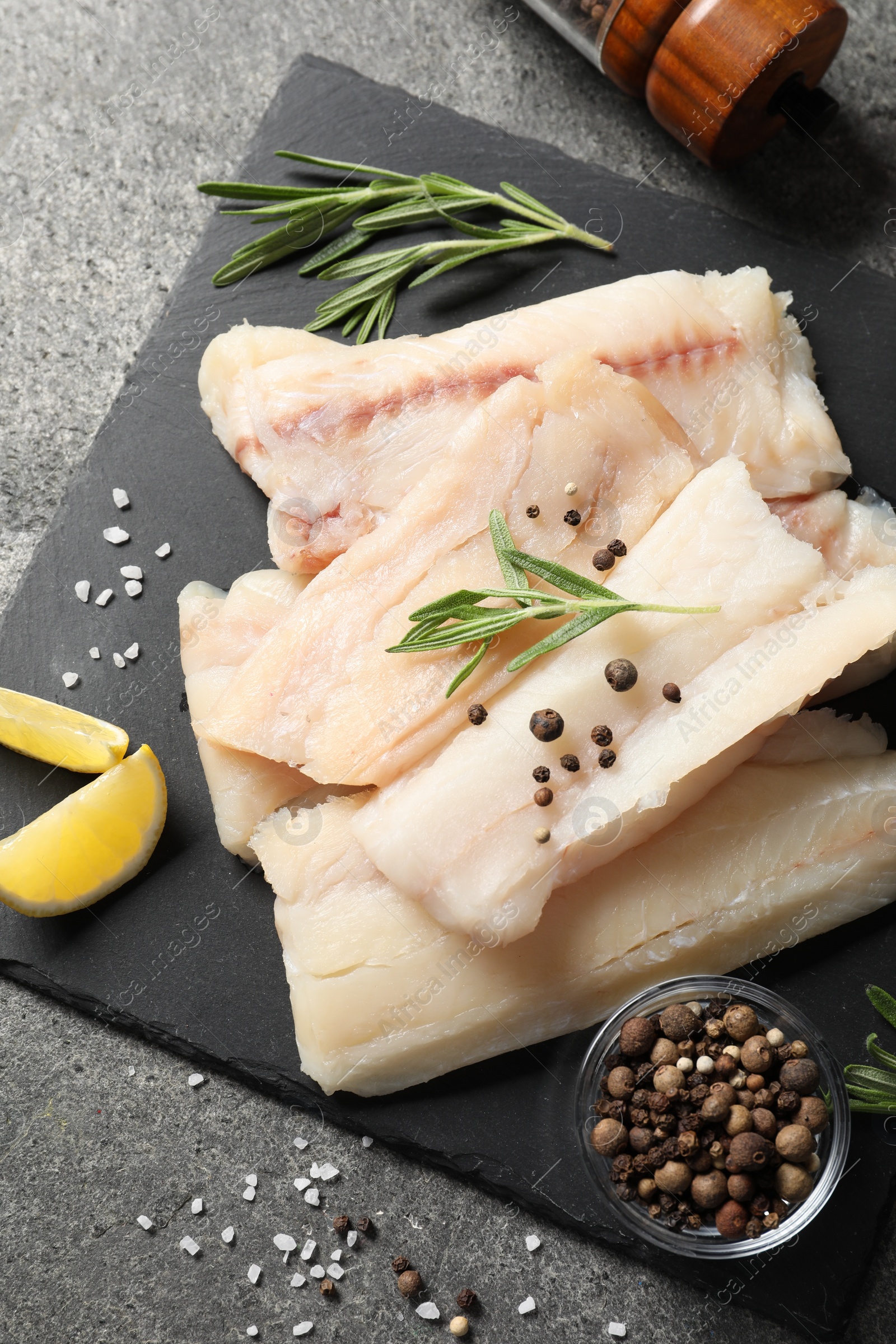 Photo of Pieces of raw cod fish, rosemary, peppercorns and lemon on grey table, top view