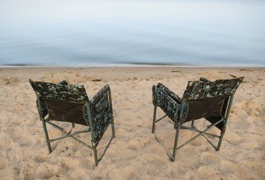 Photo of Camouflage fishing chairs on sandy beach near river