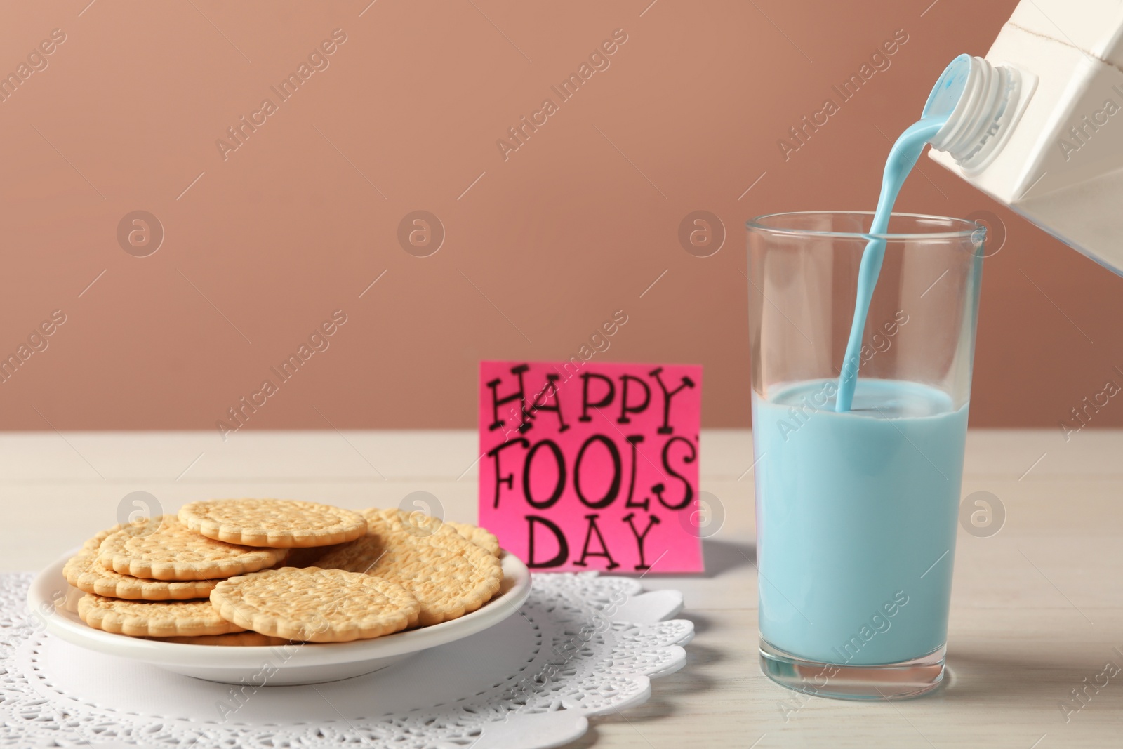 Photo of Pouring light blue milk into glass, cookies and words Happy Fool's Day on table