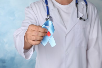 Photo of Doctor holding light blue ribbon with paper blood drop as diabetes awareness symbol on color background, closeup