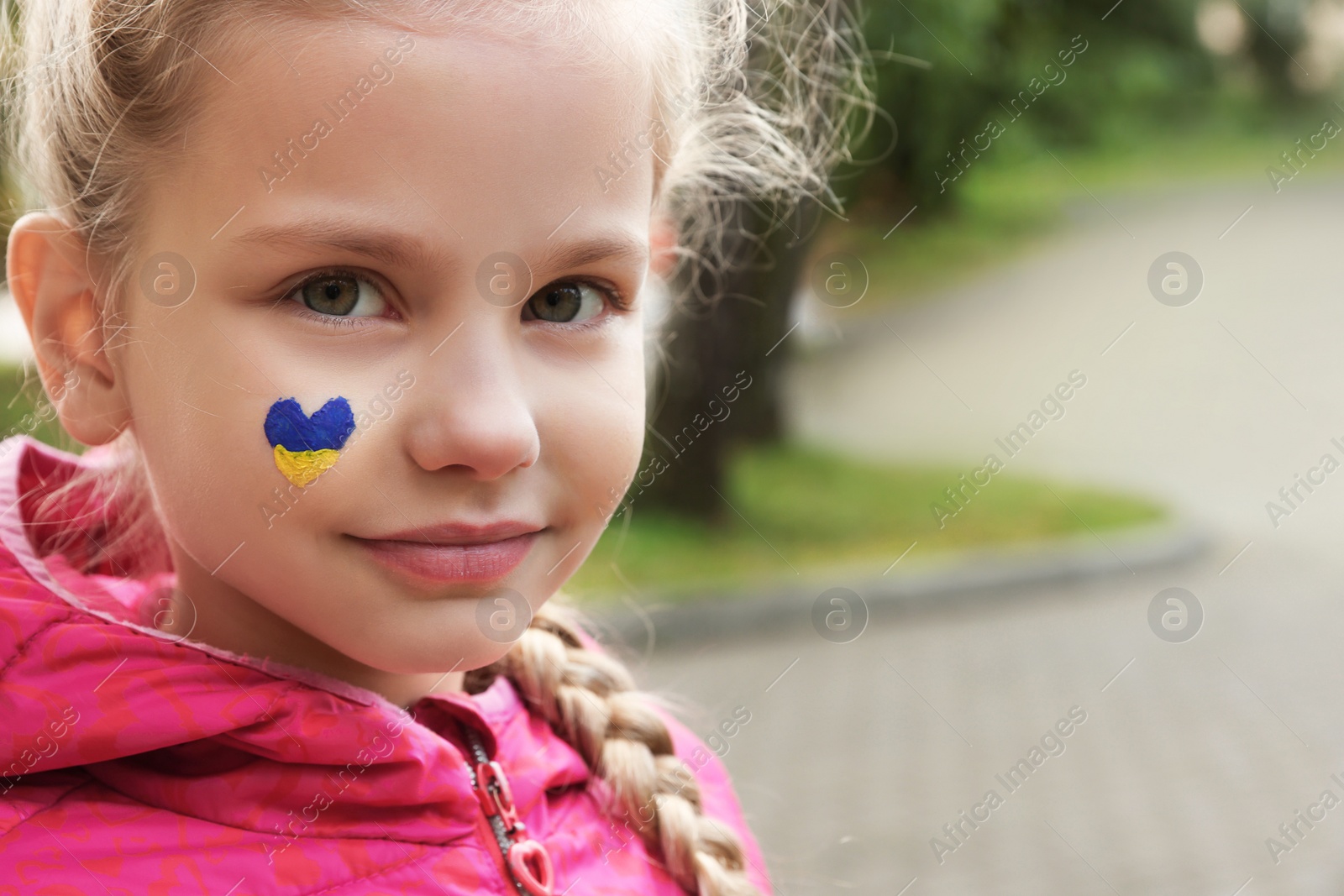 Photo of Little girl with drawing of Ukrainian flag on face in heart shape outdoors, space for text