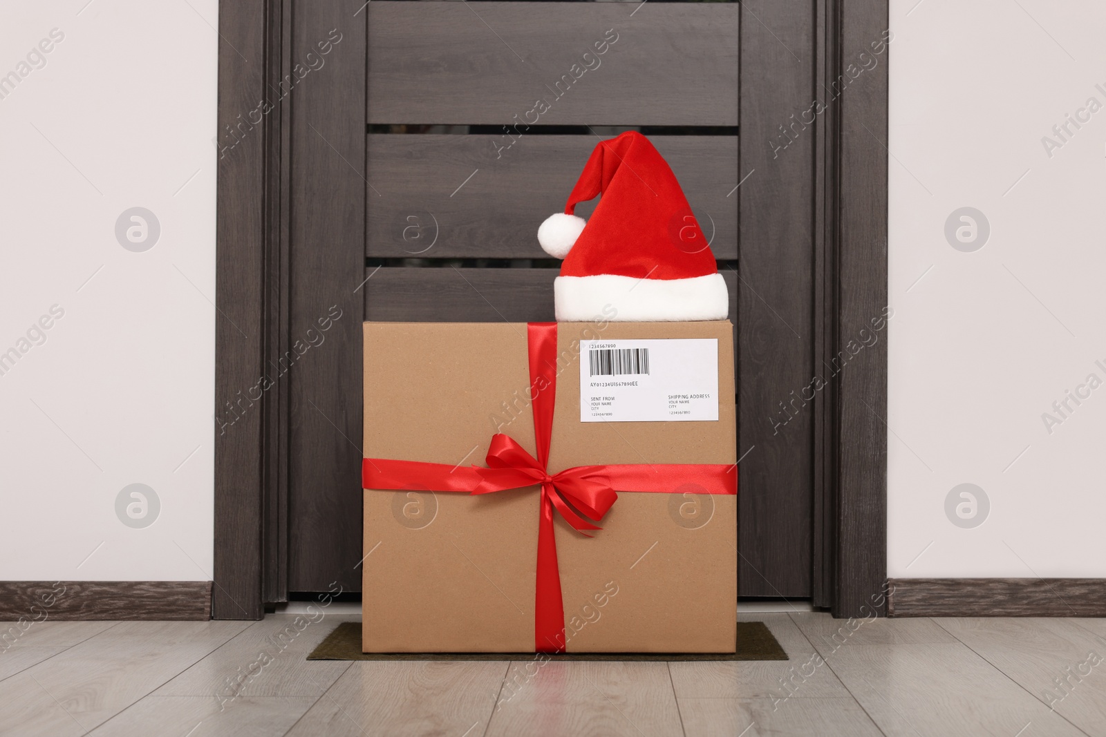 Photo of Christmas gift box and Santa hat near door indoors. Sending present by mail