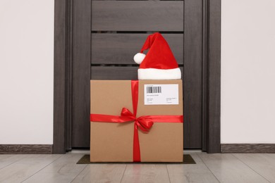 Photo of Christmas gift box and Santa hat near door indoors. Sending present by mail