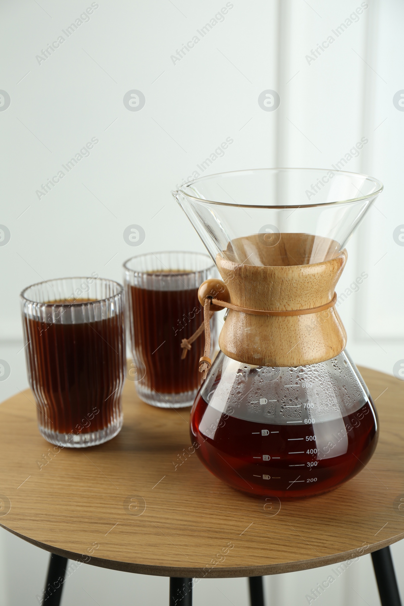 Photo of Glass chemex coffeemaker and glasses of coffee on wooden table against white wall