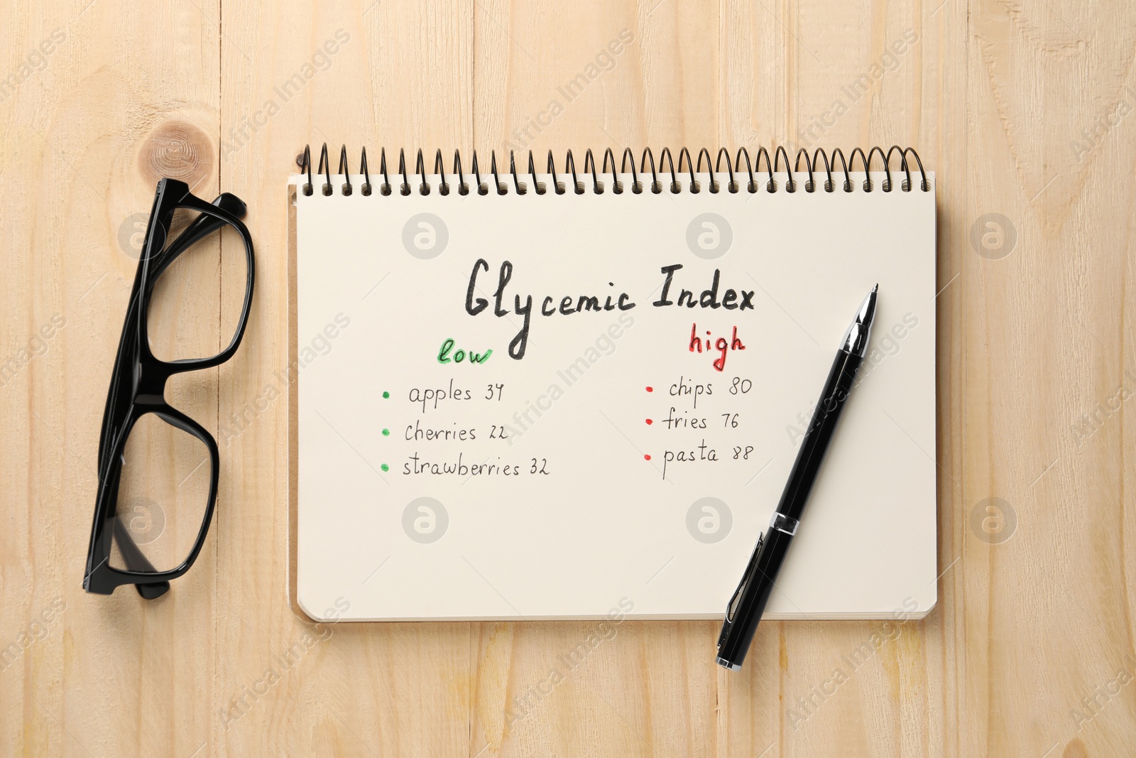 Photo of List with products of low and high glycemic index in notebook, glasses and pen on wooden table, flat lay