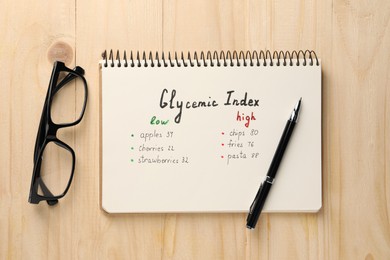 Photo of List with products of low and high glycemic index in notebook, glasses and pen on wooden table, flat lay