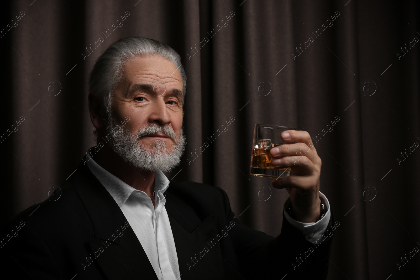 Photo of Senior man in formal suit holding glass of whiskey with ice cubes on brown background