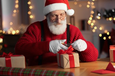 Photo of Santa Claus tying bow on gift box at his workplace in room decorated for Christmas