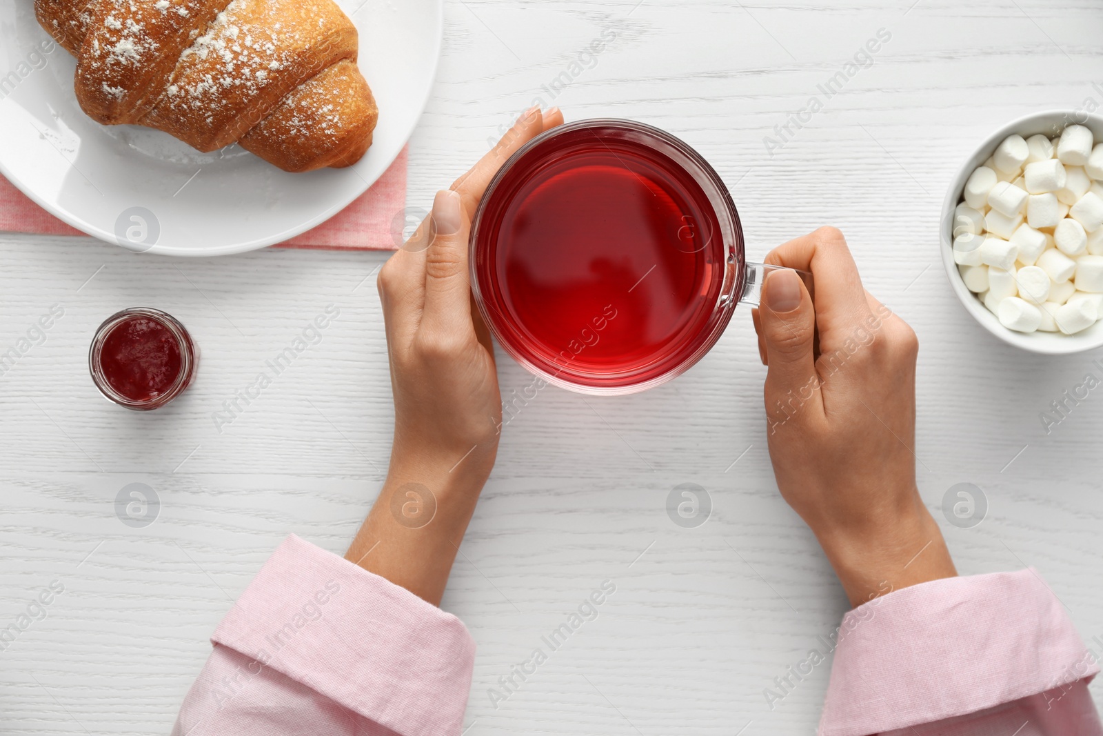 Photo of Woman holding glass cup of raspberry tea at white wooden table, top view. Delicious morning meal