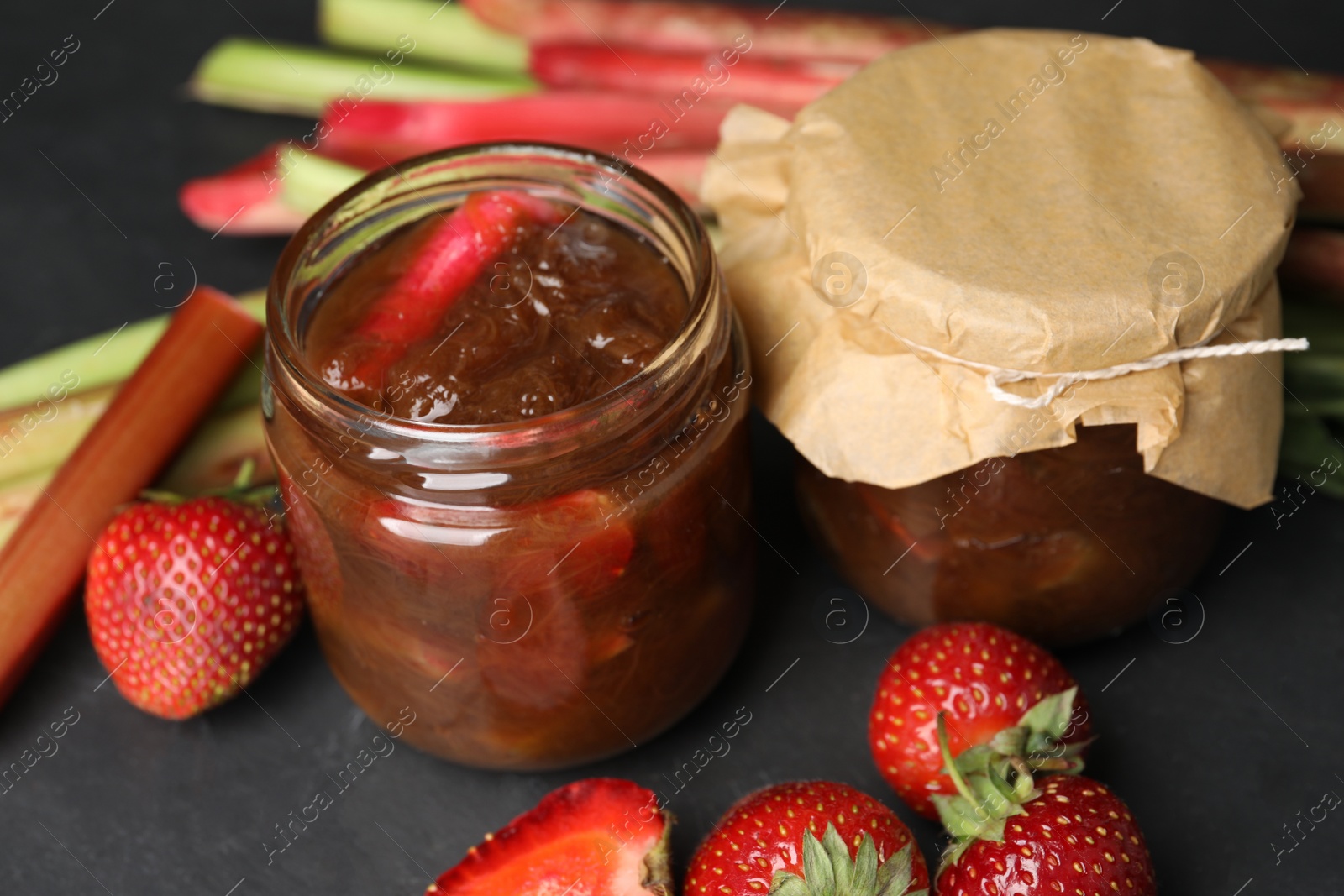 Photo of Jars of tasty rhubarb jam, fresh stems and strawberries on dark textured table, closeup