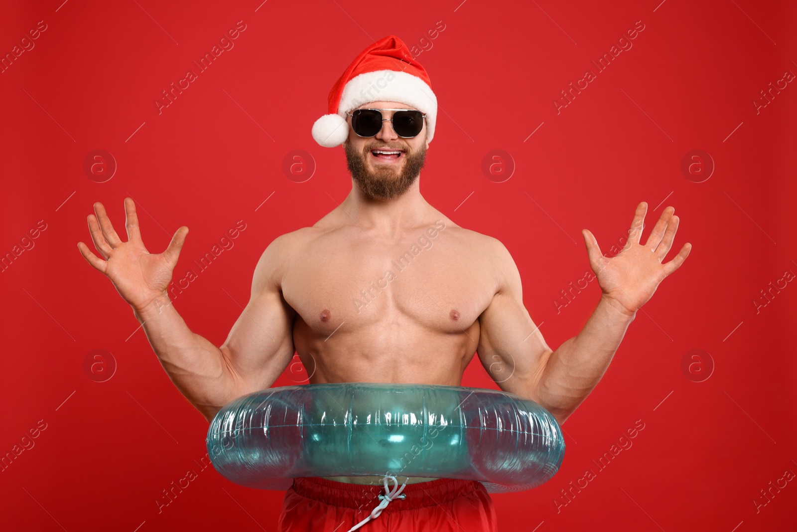 Photo of Muscular young man in Santa hat and sunglasses with inflatable ring on red background