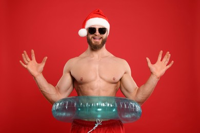 Muscular young man in Santa hat and sunglasses with inflatable ring on red background