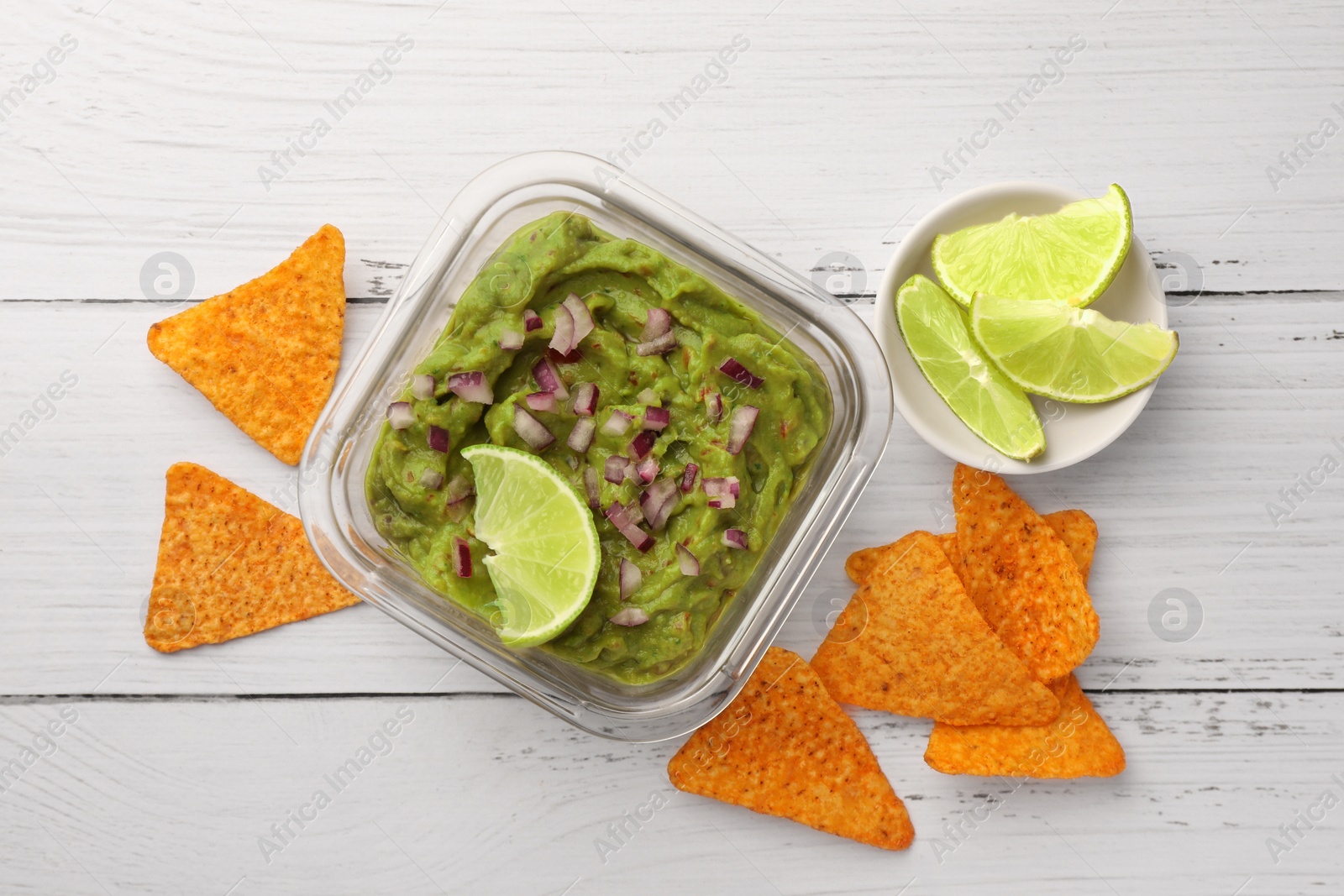 Photo of Bowl of delicious guacamole with onion, lime and nachos chips on white wooden table, flat lay