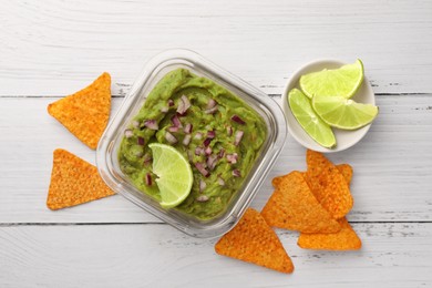 Photo of Bowl of delicious guacamole with onion, lime and nachos chips on white wooden table, flat lay