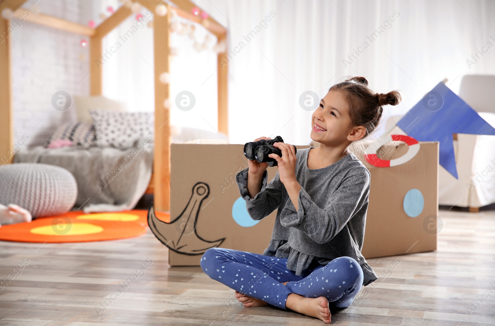 Photo of Cute little girl playing with binoculars and cardboard boat in bedroom