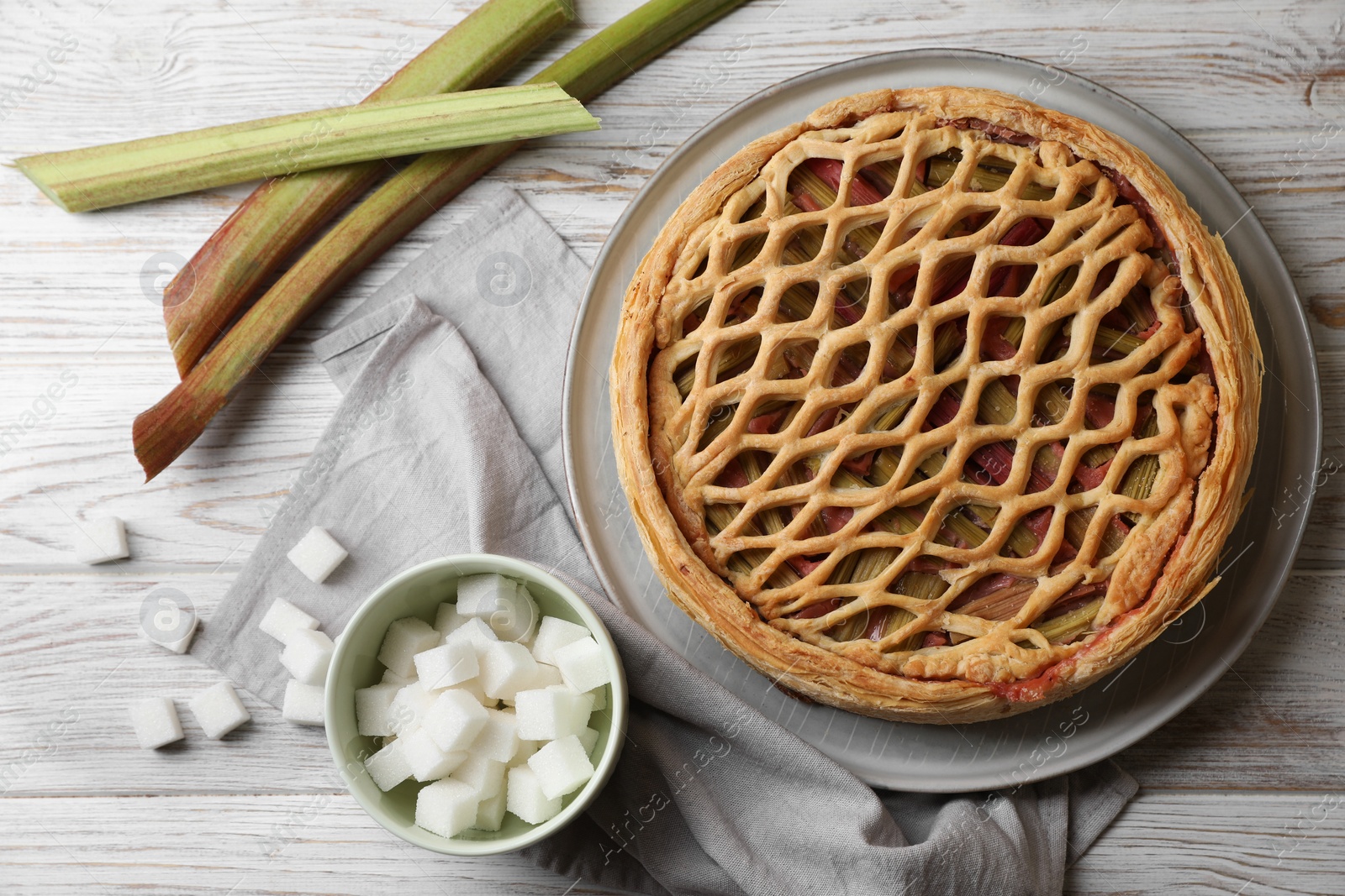 Photo of Freshly baked rhubarb pie, stalks and sugar cubes on light wooden table, flat lay