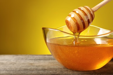 Delicious honey flowing down from dipper into bowl on wooden table against yellow background, closeup. Space for text