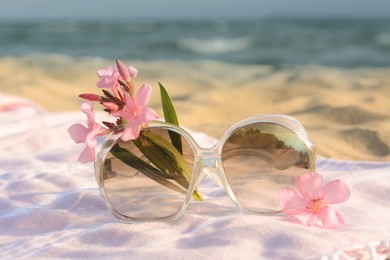 Photo of Beautiful sunglasses with tropical flower on blanket near sea, closeup