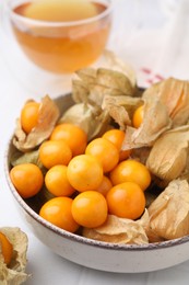 Ripe physalis fruits with calyxes in bowl on white table, closeup