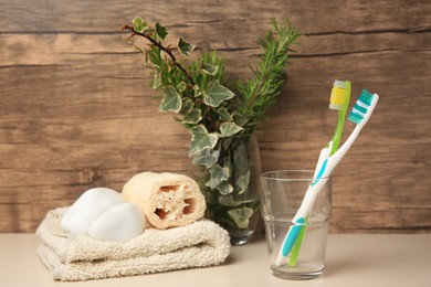 Photo of Toothbrushes in glass, towel, loofah and stones on beige table