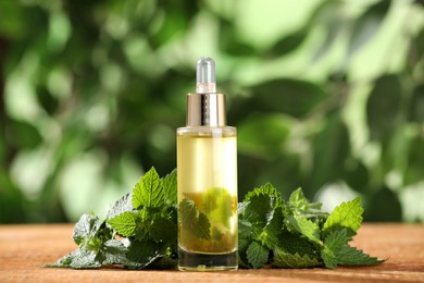 Photo of Glass bottle of nettle oil with dropper and leaves on wooden table against blurred background
