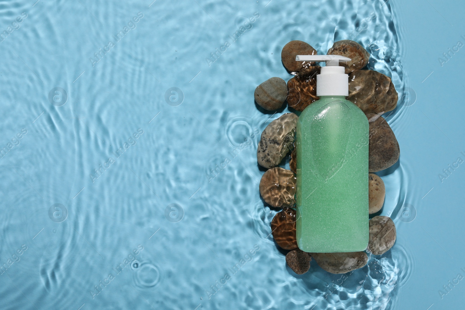 Photo of Bottle of face cleansing product and stones in water against light blue background, flat lay. Space for text