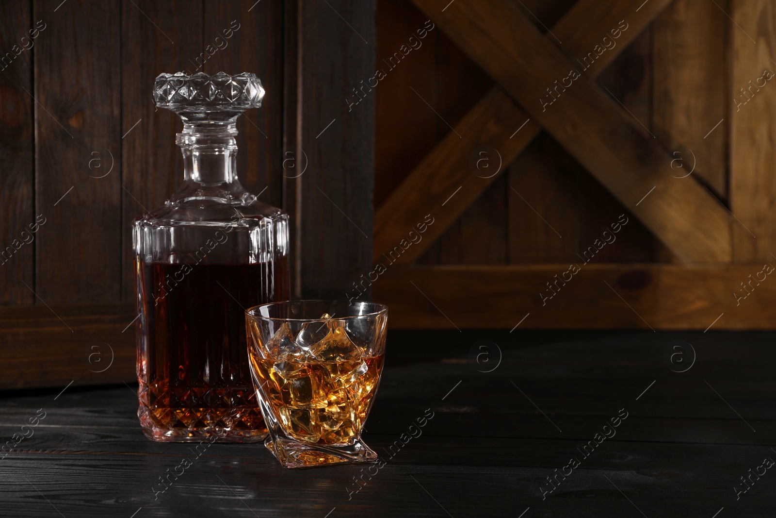 Photo of Whiskey with ice cubes in glass and bottle on black wooden table, space for text