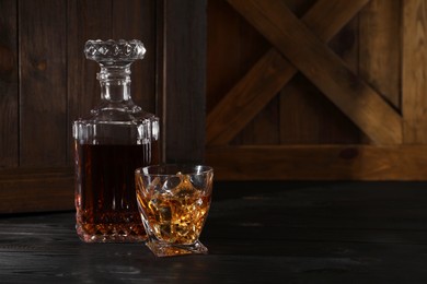 Photo of Whiskey with ice cubes in glass and bottle on black wooden table, space for text
