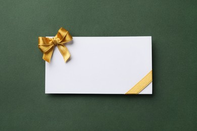 Photo of Blank gift card with golden bow on dark green background, top view