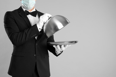 Photo of Waiter in medical face mask holding tray with lid on light grey background, closeup. Space for text