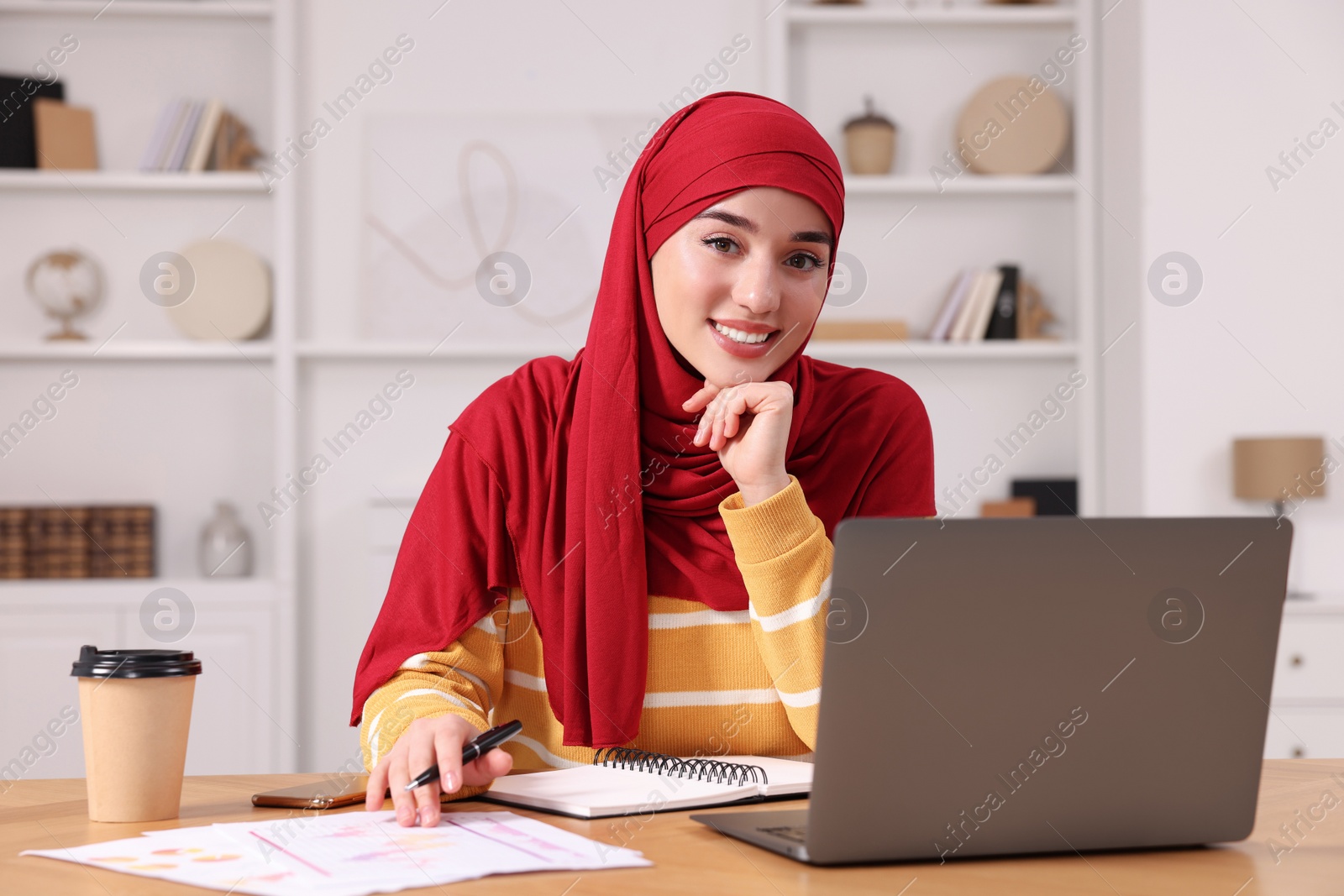 Photo of Muslim woman in hijab using laptop at wooden table in room