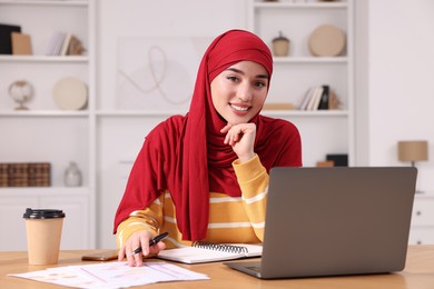 Muslim woman in hijab using laptop at wooden table in room