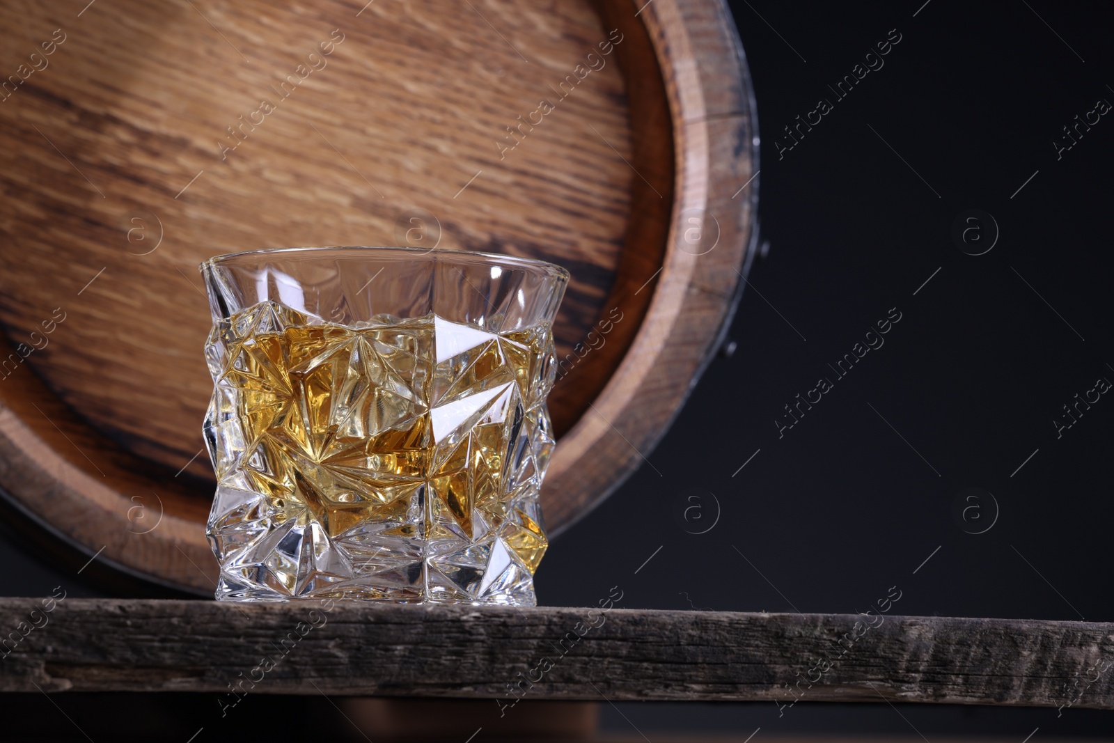 Photo of Whiskey with ice cubes in glass and barrel on wooden table against black background, closeup. Space for text