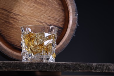Whiskey with ice cubes in glass and barrel on wooden table against black background, closeup. Space for text