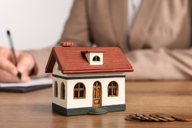 Mortgage concept. Woman writing something in notebook, house model and coins on wooden table, selective focus