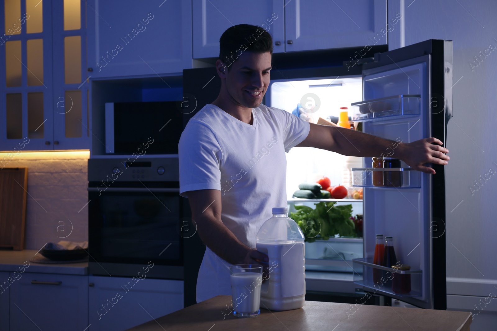 Photo of Man holding gallon bottle of milk and glass on wooden table in kitchen at night