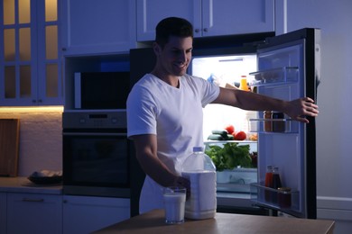 Photo of Man holding gallon bottle of milk and glass on wooden table in kitchen at night