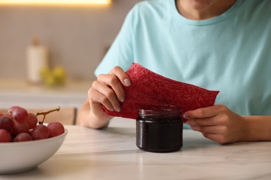 Photo of Woman packing jar of jam into beeswax food wrap at light table in kitchen, closeup