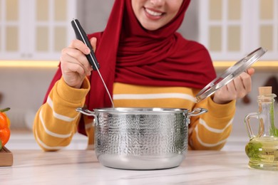 Muslim woman making delicious soup with vegetables at white table in kitchen, closeup