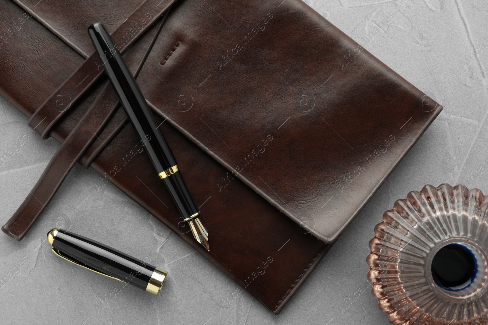 Photo of Stylish fountain pen, leather notebook and inkwell on light grey textured table, flat lay