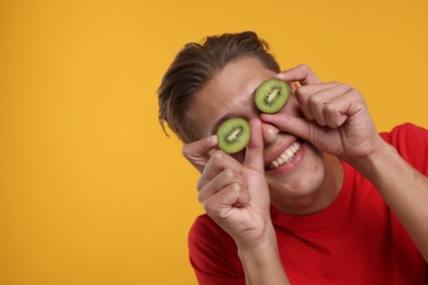 Smiling man covering eyes with halves of kiwi on orange background. Space for text