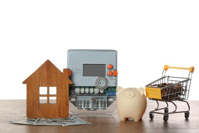 Photo of Electricity meter, house model, piggy bank and shopping cart with money on wooden table against white background