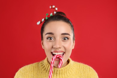 Photo of Surprised young woman in yellow sweater and festive headband eating candy cane on red background, closeup