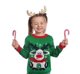 Photo of Cute little girl in Christmas sweater and deer headband holding candy canes on white background