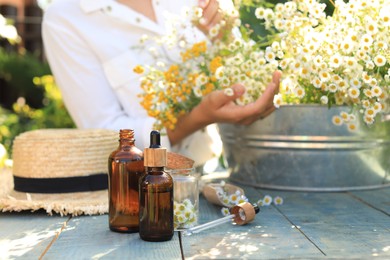 Woman with flowers near table outdoors, focus on bottles of chamomile essential oil