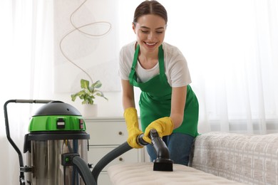 Photo of Professional janitor in uniform vacuuming furniture indoors