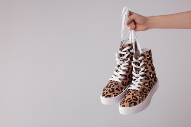 Photo of Woman holding pair of sneakers with leopard print on light grey background, closeup. Space for text