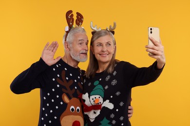 Photo of Senior couple in Christmas sweaters and reindeer headbands taking selfie on orange background