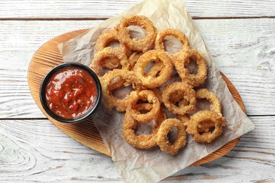 Photo of Homemade crunchy fried onion rings with tomato sauce on wooden table, top view
