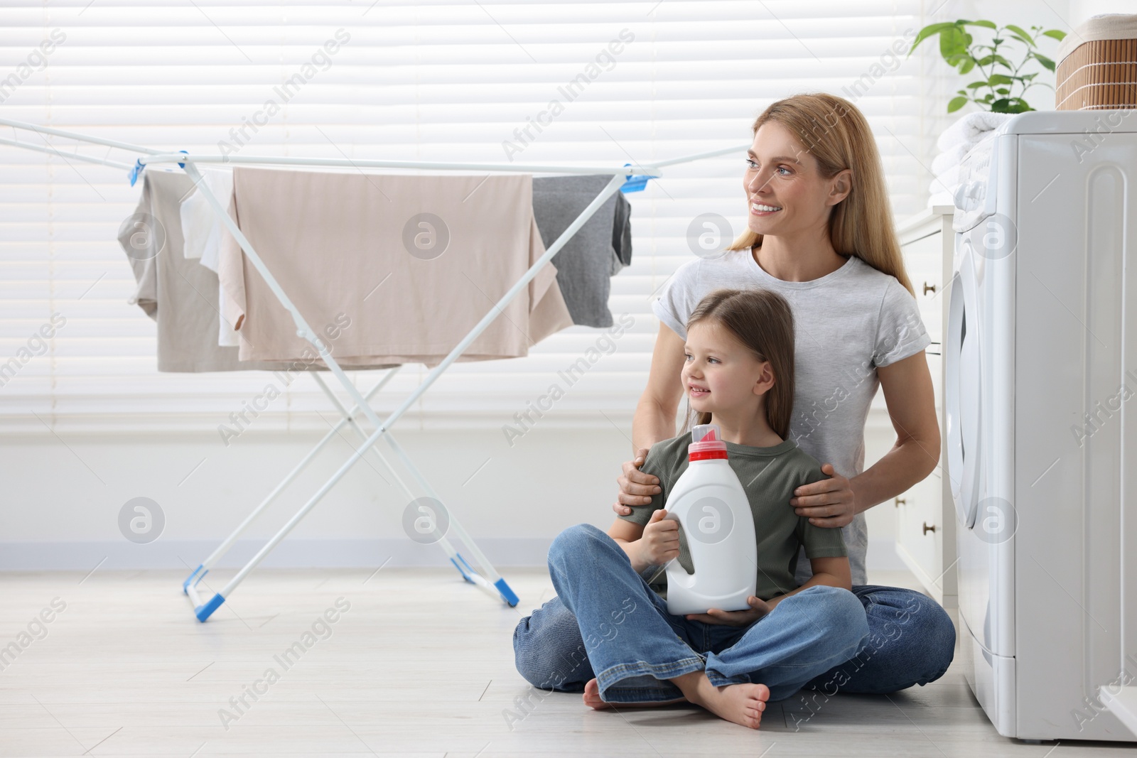 Photo of Mother and daughter sitting on floor with fabric softener near clothes dryer in bathroom, space for text